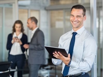 Happy businessman holding digital tablet with team discussing project in the background. Portrait of a confident business man holding computer laptop. Successful young business man at the office.